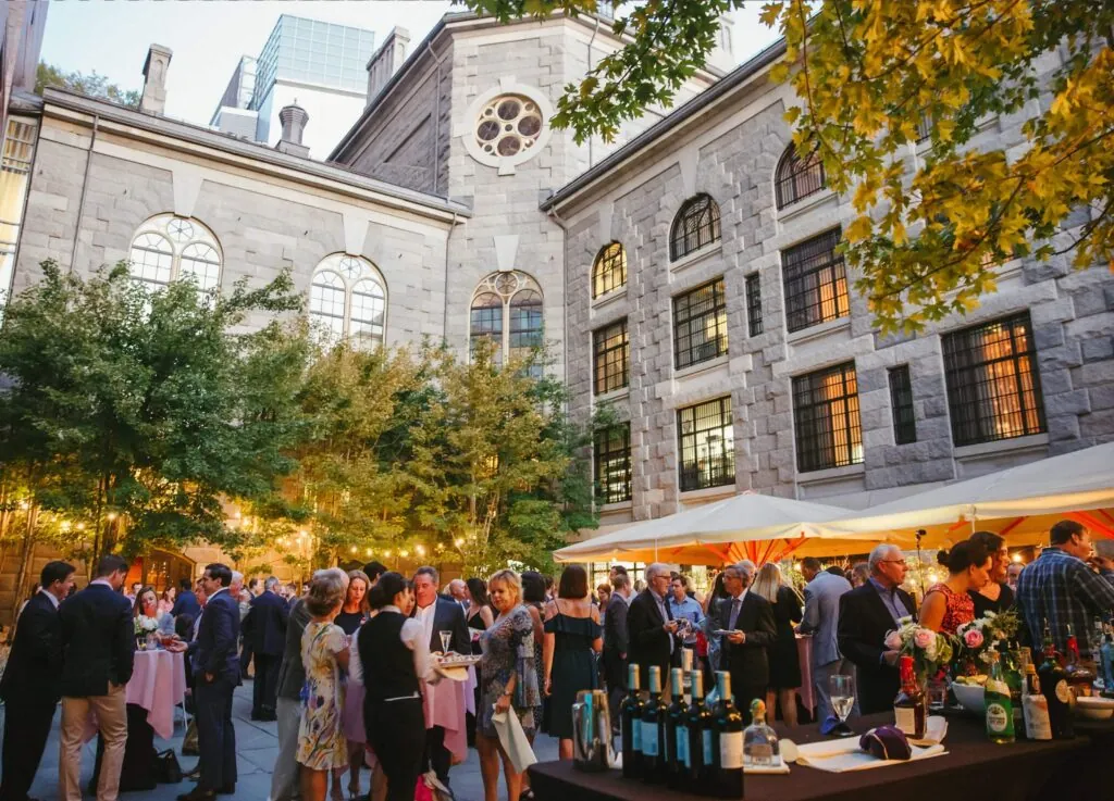 People socialize at an outdoor evening event with lights, drinks, and food in a historic stone courtyard at a boutique hotel surrounded by trees.