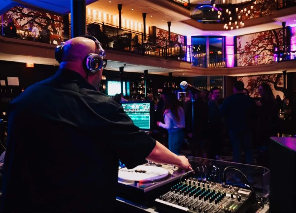 A DJ wearing headphones mixes music at a lively hotel event with people socializing, colorful lighting, and a stylish, gallery-inspired venue.