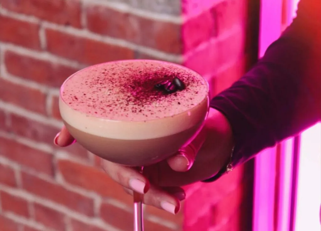 A photo of a hand holding a creamy espresso martini in a coupe glass, topped with coffee beans, against a brick wall in hotel-worthy pink lighting.