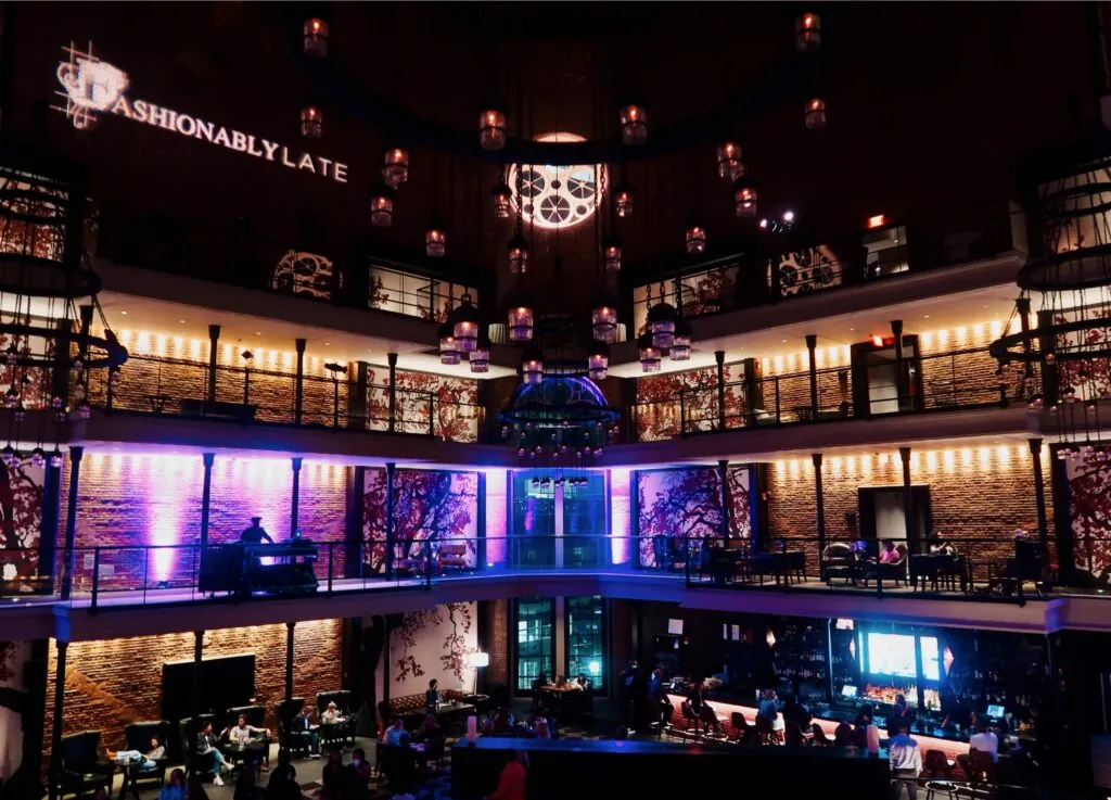 A stylish, dimly lit Beacon Hill lounge with dining, live piano, purple lighting, and FASHIONABLY LATE projected on the wall—perfect for a photo.