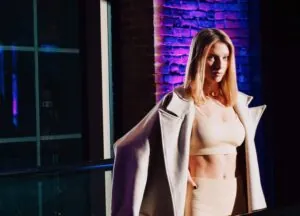 A woman in a beige crop top and skirt with a light coat poses for a luxury photo indoors, set against a purple-lit brick wall in Beacon Hill.