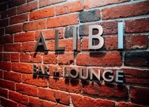 Metal letters spelling ALIBI BAR + LOUNGE glow with warm lighting on a red brick wall, adding a touch of luxury to the hotel’s vibrant ambiance.