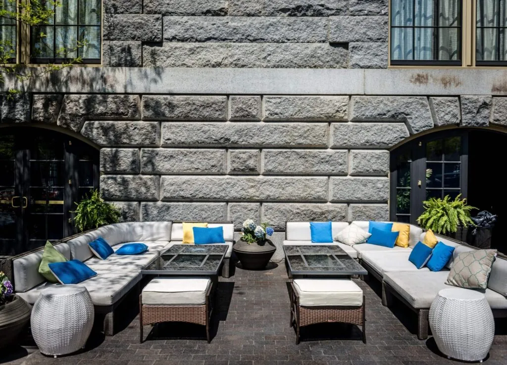 Photo of an outdoor patio with L-shaped sofas, bright pillows, glass-top tables, and potted plants—perfect for a Beacon Hill gallery.