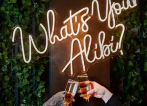 Two hands clink champagne glasses in front of a neon sign at a chic Beacon Hill hotel, with green leaves and style that’s pure gallery cool.