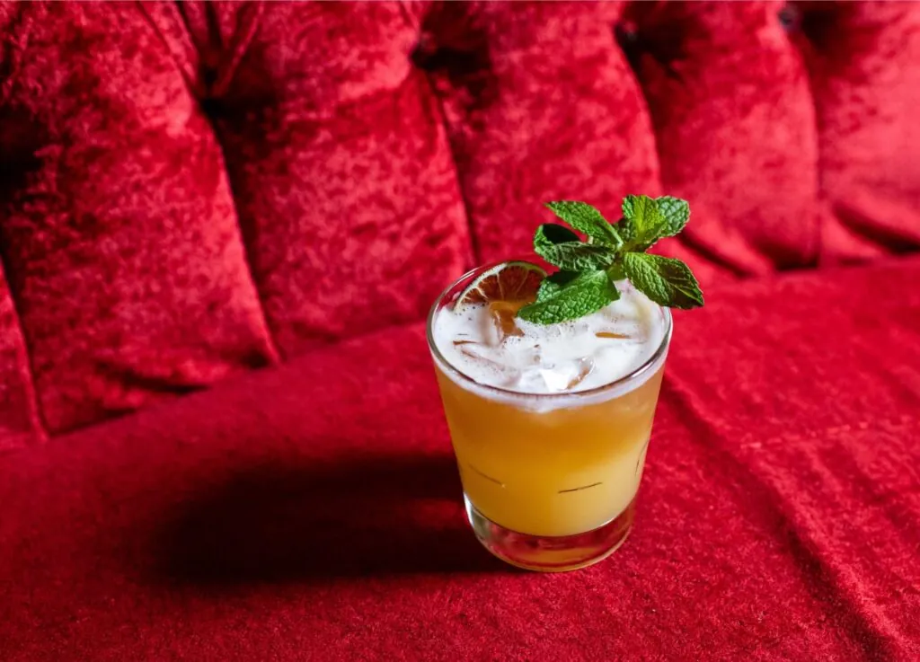 A yellow cocktail with ice and a mint garnish sits on a plush red velvet surface, exuding luxury worthy of a five-star hotel.