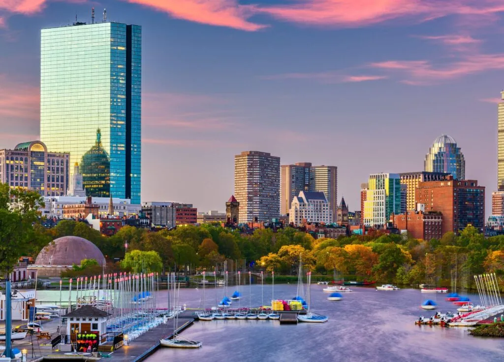 Luxury hotel views of the Boston skyline at sunset with sailboats on the Charles River, skyscraper reflections, and colorful clouds.