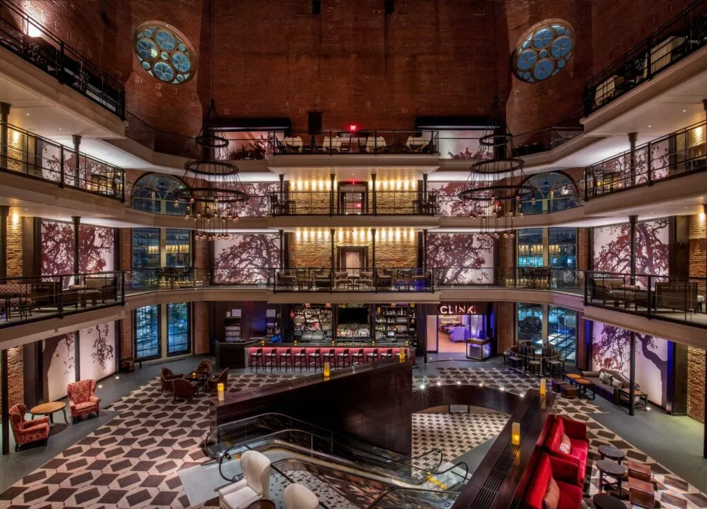 A spacious, luxury hotel lobby with balconies, a central staircase, bar area, modern lighting, patterned floor, and tall decorative walls.