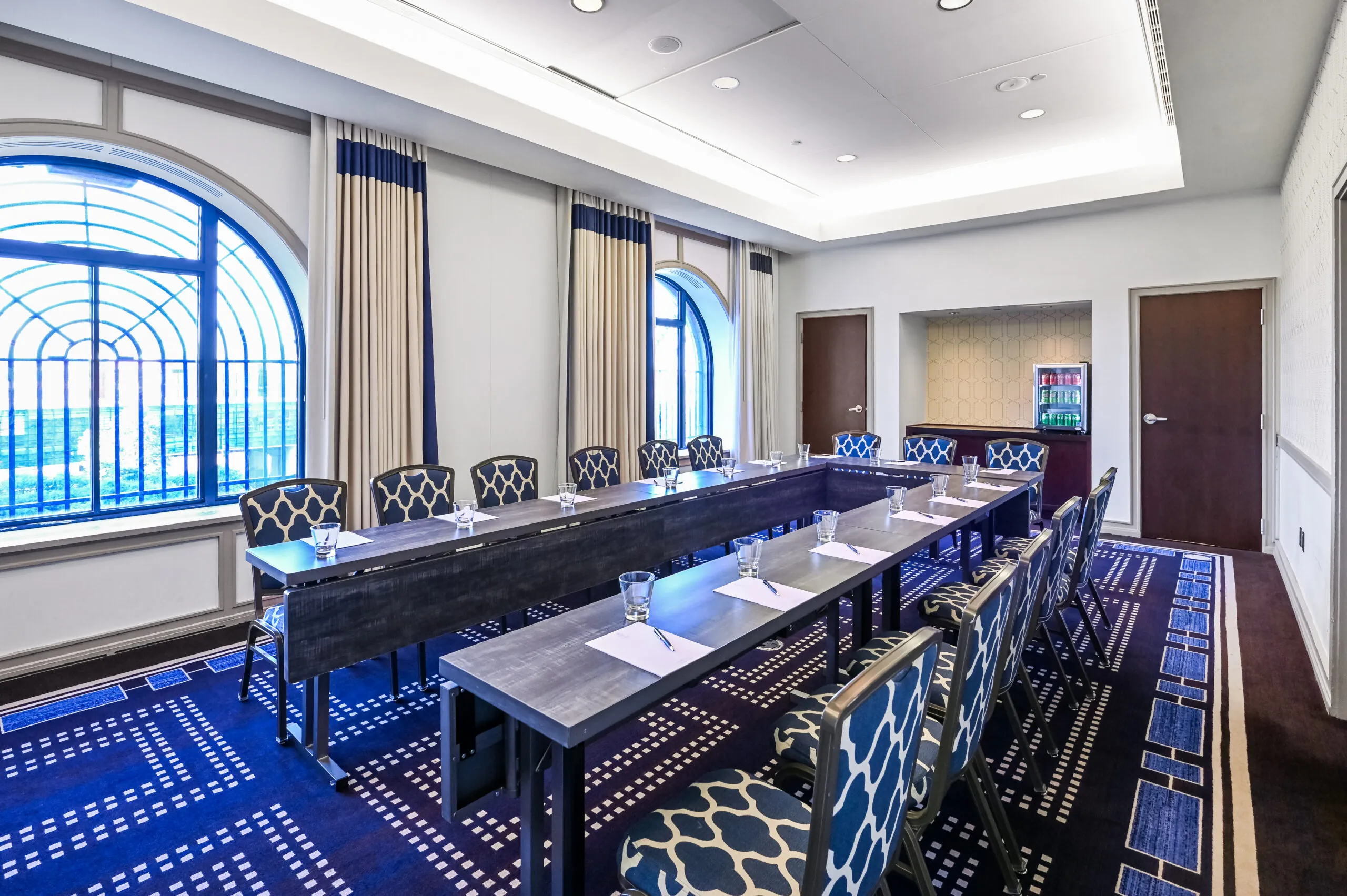 Conference room meeting space with U-shaped table, blue chairs, notepads, glasses, and large arched windows overlooking Boston Common.