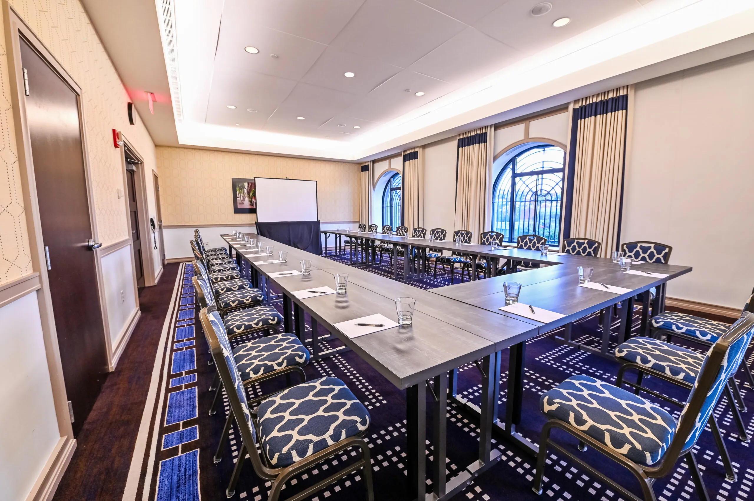Modern Boston conference space with U-shaped table, patterned chairs, notepads, water glasses, large windows, and projection screen near Boston Common.