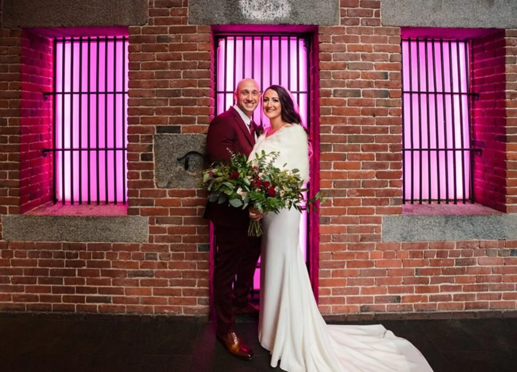 A bride and groom smile in front of glowing pink windows at a hotel, holding a bouquet and dressed in formal wedding attire for their photo.
