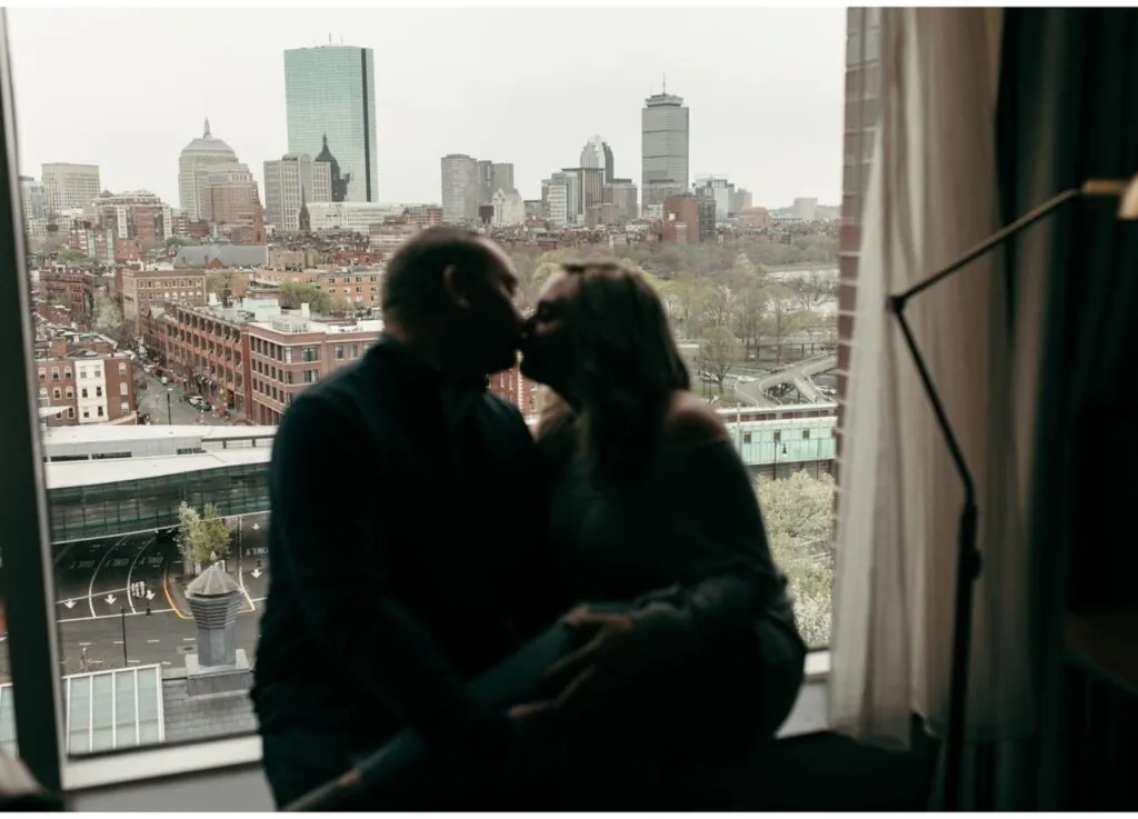 A couple kisses on a hotel windowsill, silhouetted against a cityscape of tall buildings and overcast skies in this romantic photo.