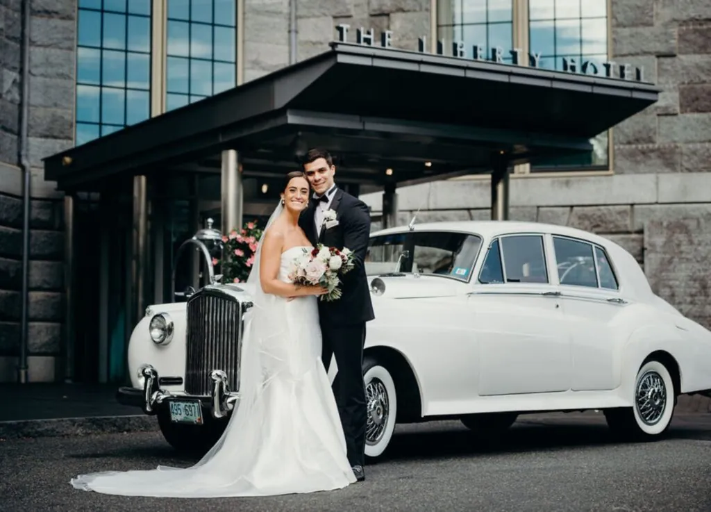 A bride and groom in wedding attire pose with a bouquet by a classic white car outside the luxury Liberty Hotel.
