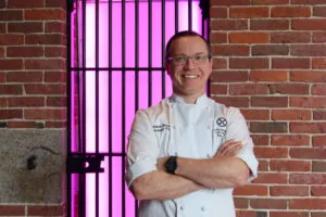 Smiling chef in a white uniform stands with arms crossed in front of a brick wall and bars, with a bright pink light in the background.