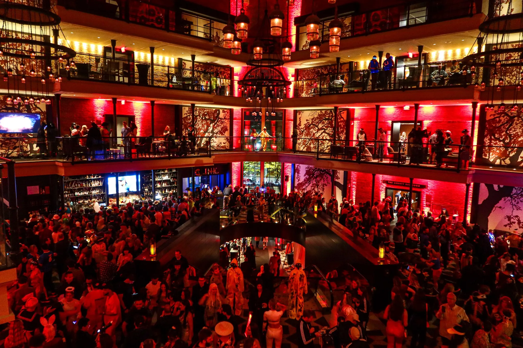 Crowded, vibrant indoor party with multiple balconies, red lighting, and people in costumes mingling across three floors.