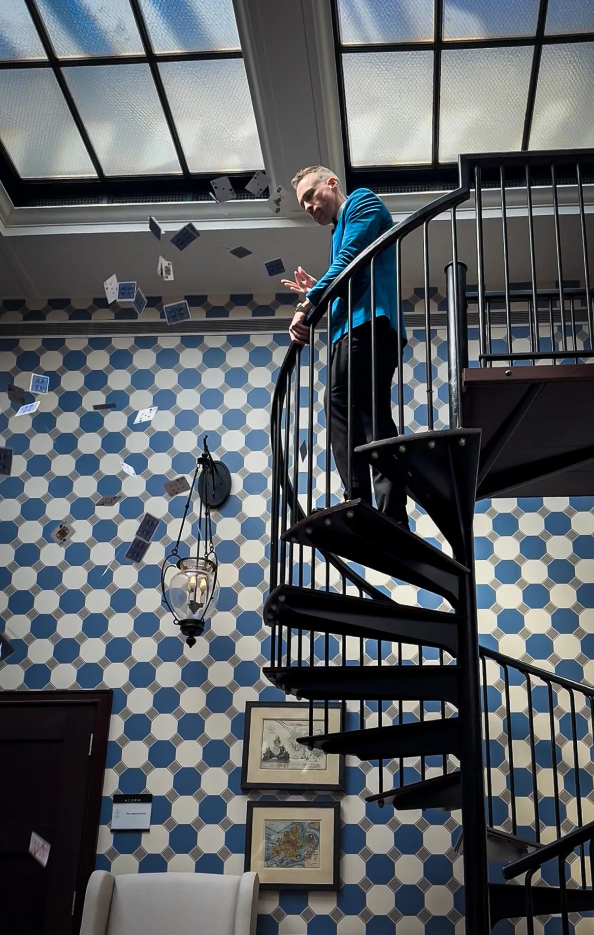 A man in a blue jacket stands on a spiral staircase, tossing playing cards into the air in a room with patterned walls and framed pictures.