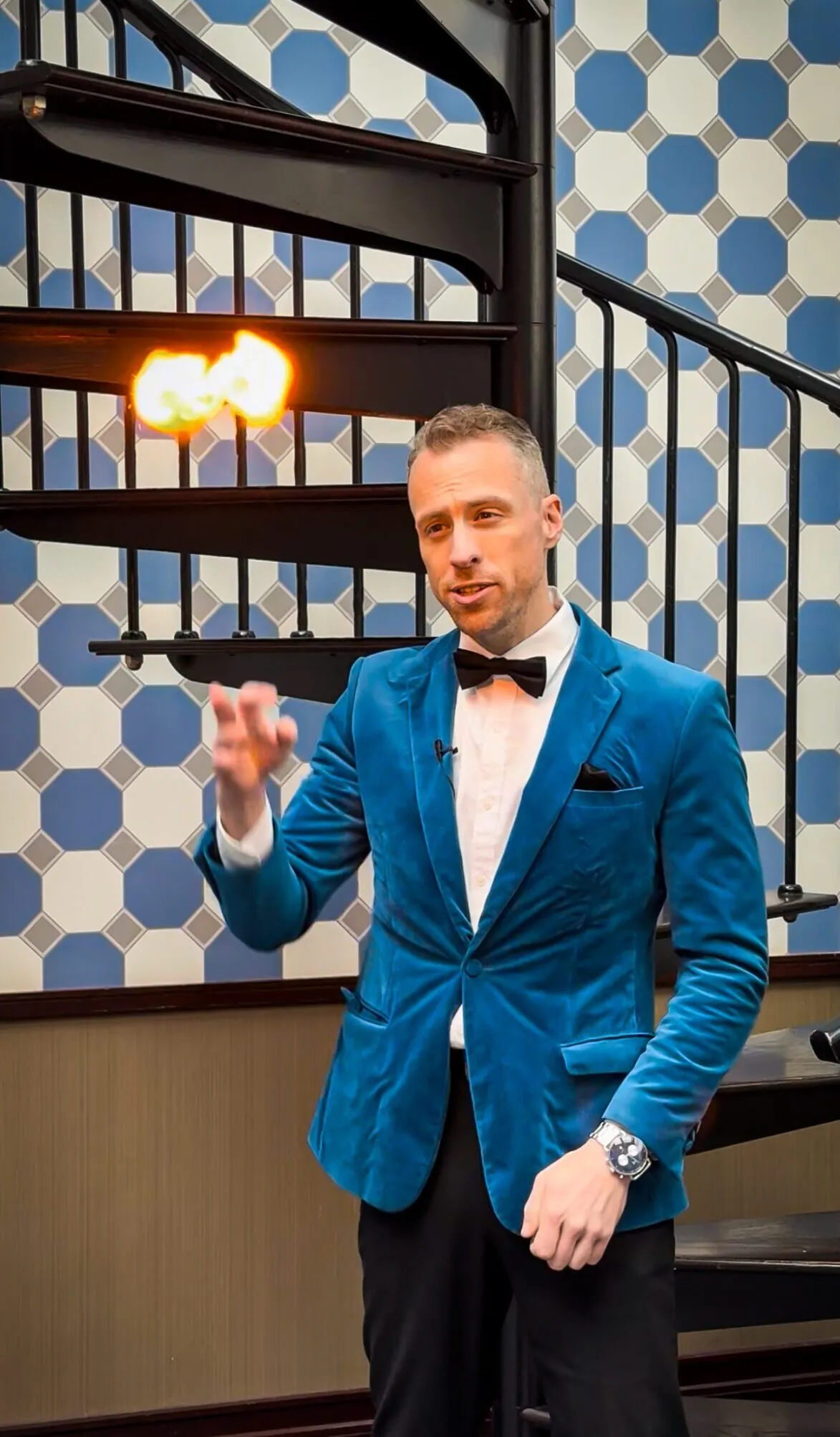 A man in a blue velvet suit jacket and bow tie performs a fire trick, holding a small flame in the air by a spiral staircase.