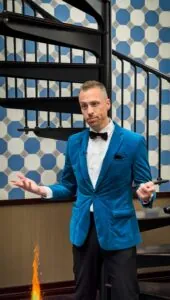 A man in a blue velvet blazer and bow tie stands on a spiral staircase, gesturing with his hands. There is a small flame near the ground.