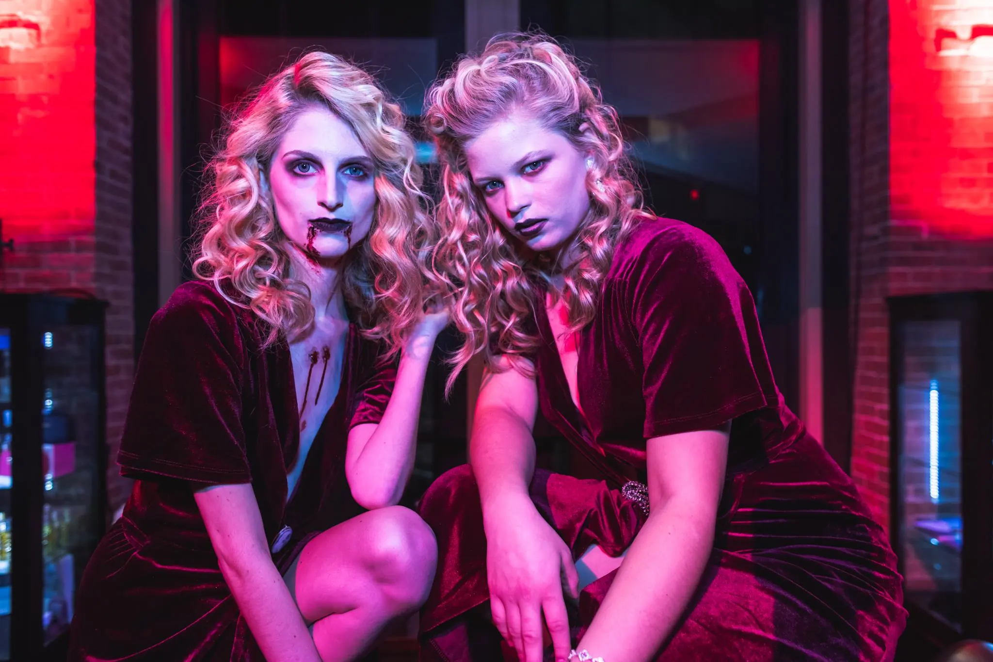Two women in red velvet dresses with curly blonde hair and dramatic makeup pose in a dimly lit room with red and purple lighting.