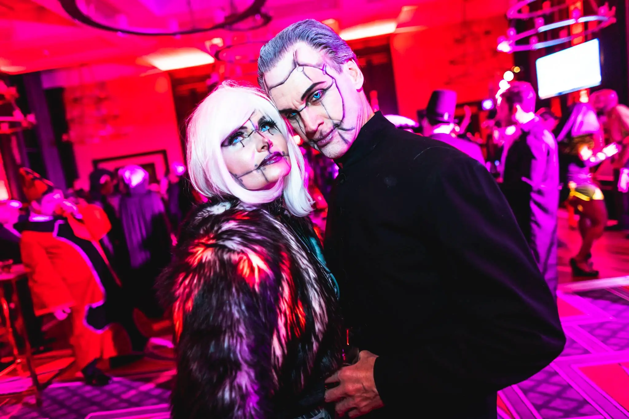 Two people in dramatic, cracked doll makeup and costumes pose together at a vibrant, red-lit costume party with other guests in the background.
