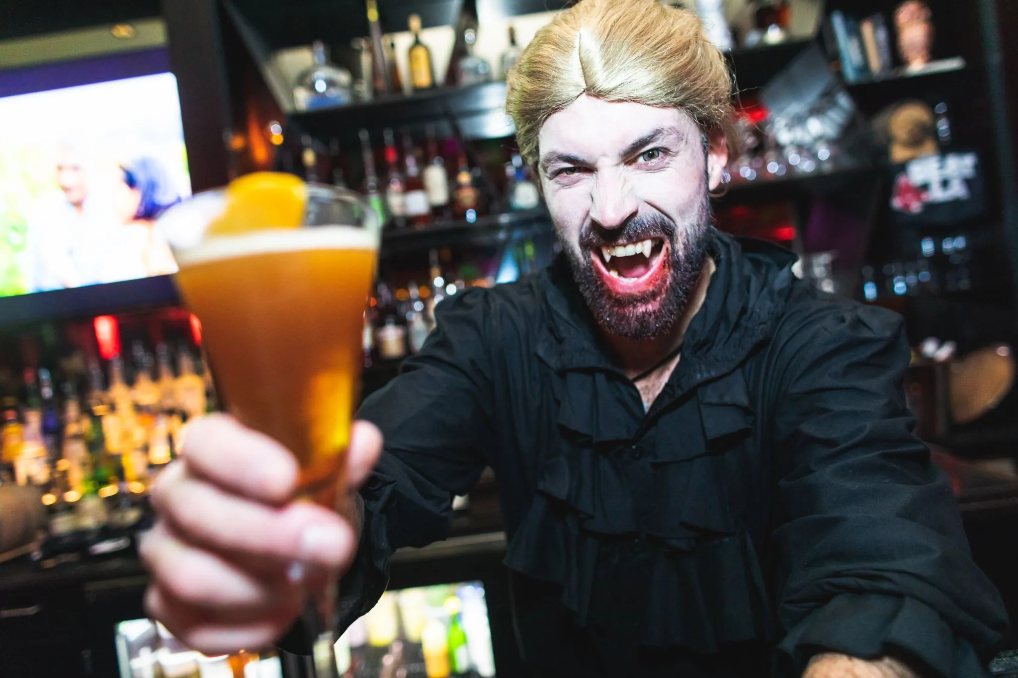 A man in vampire makeup and costume snarls while holding a cocktail in a bar, with bottles and glasses visible in the background.