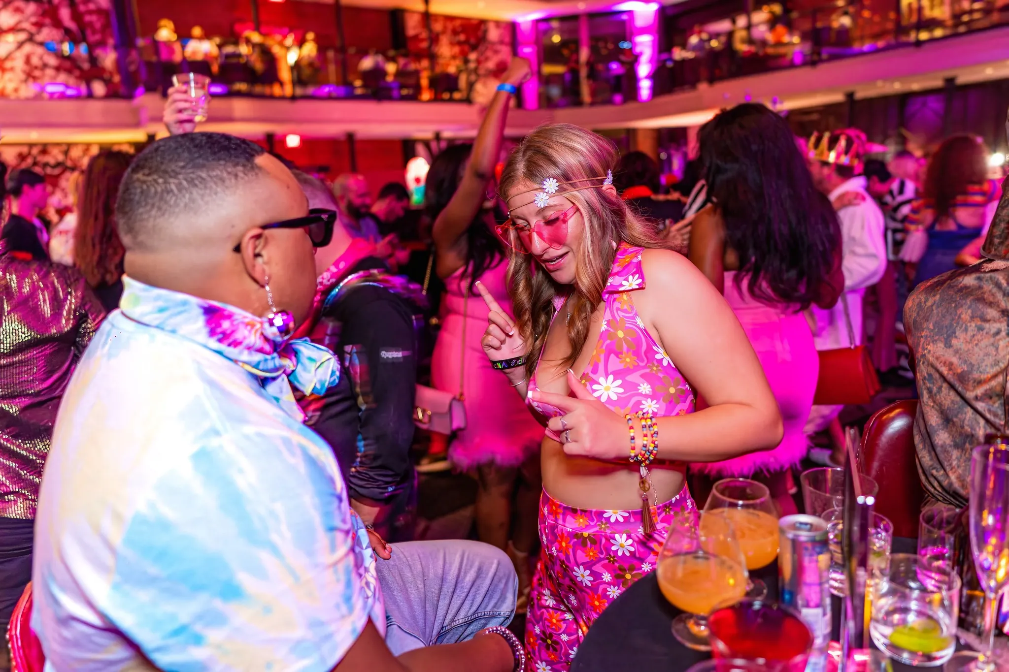Two people in colorful, 70s-inspired outfits dance and have fun at a lively, crowded party with bright, vibrant lighting.