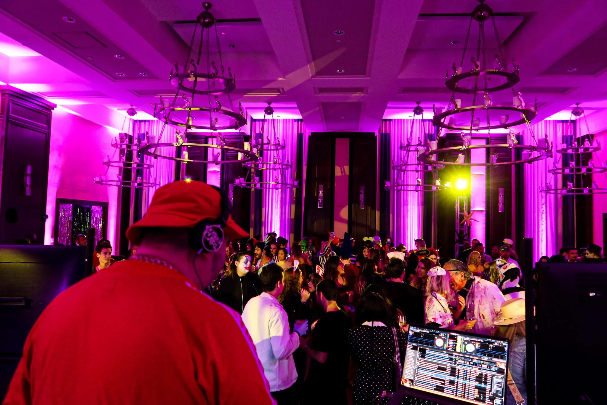A DJ in a red cap plays music for a lively crowd at a party in a purple-lit room with chandeliers.