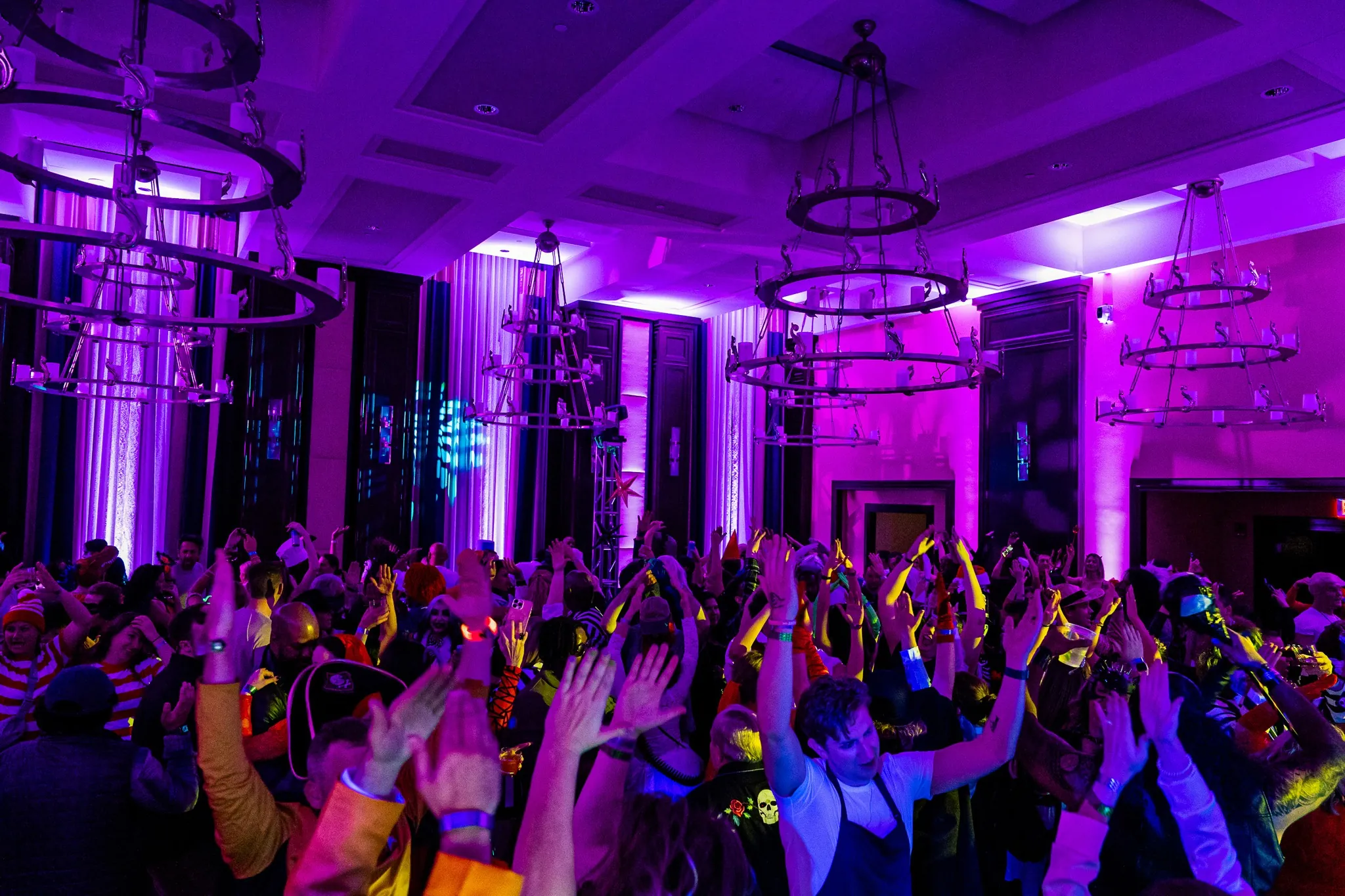 Large crowd dancing with raised hands in a colorful, purple-lit room decorated with chandeliers during a lively indoor party or event.