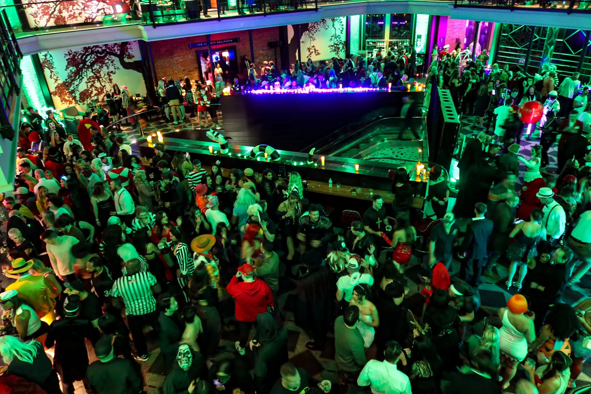 A large crowd of people in costumes gather at a lively indoor party with colorful lighting and decorations.