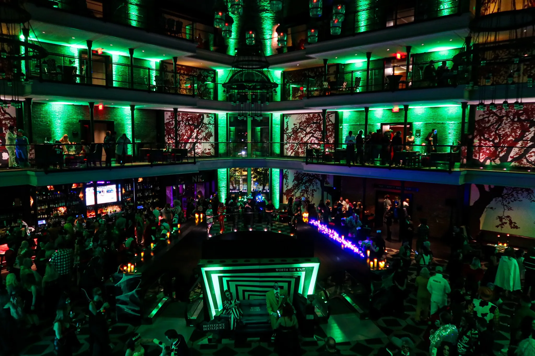 A large, multi-level indoor venue with green lighting, balconies, and a crowd of people socializing at a lively nighttime event.