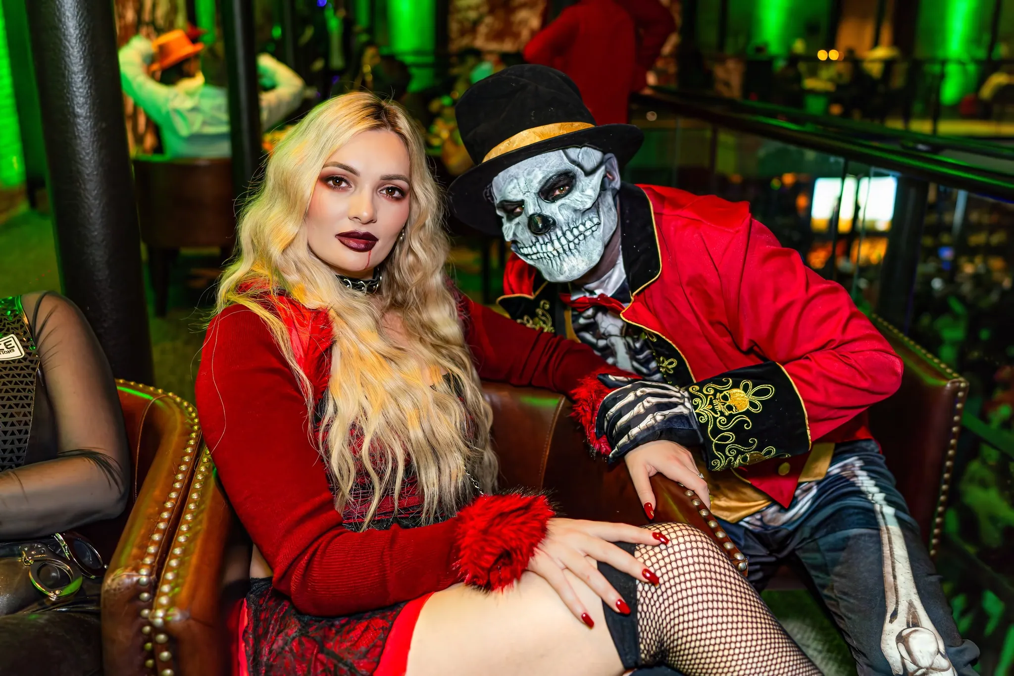 A woman in a red dress sits next to a person in a skeleton mask and red jacket at a festive, dimly lit party with green lighting.