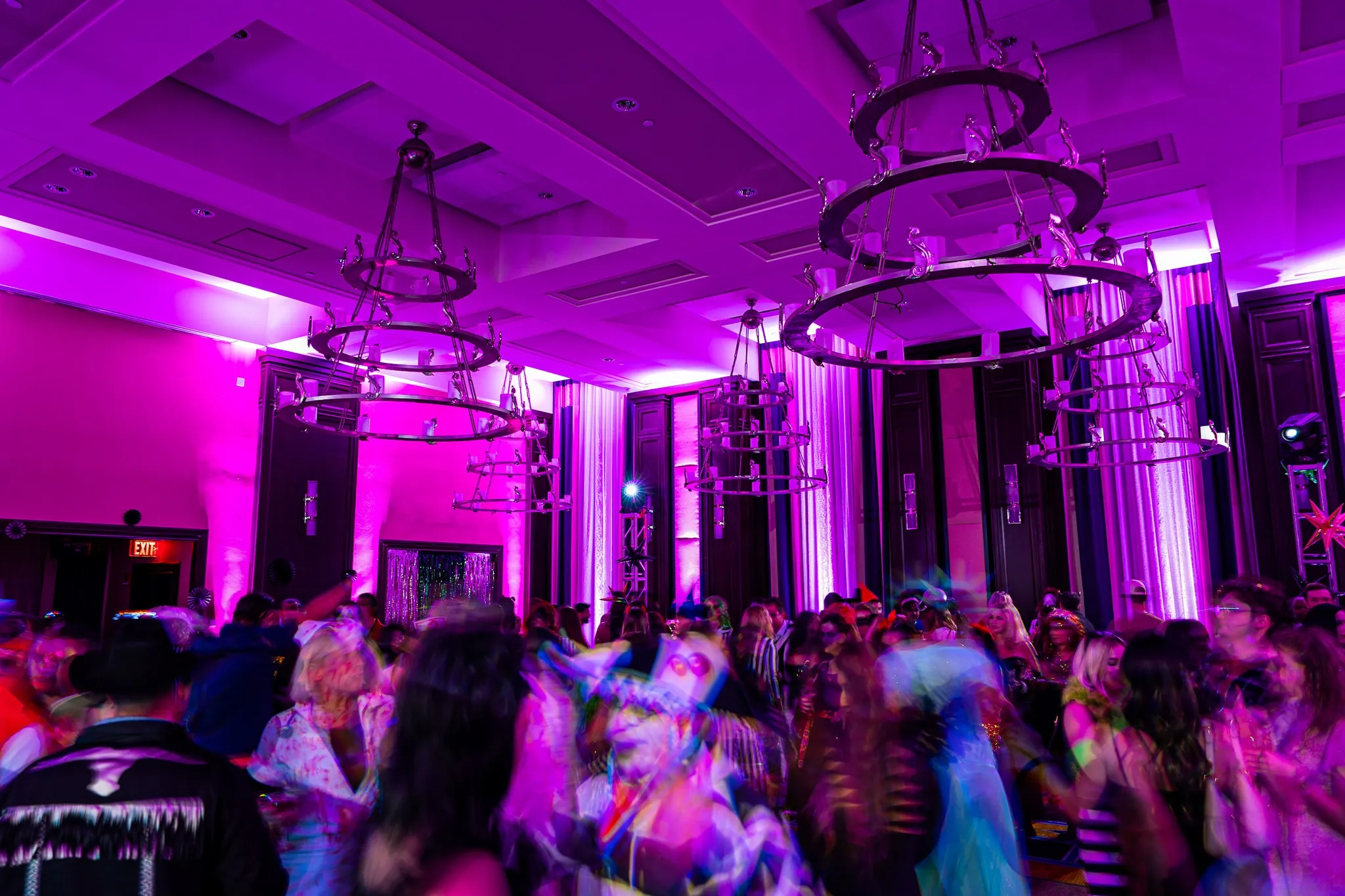 A crowd of people dance in a room with purple lighting, large chandeliers, and high ceilings, creating a vibrant, energetic atmosphere.