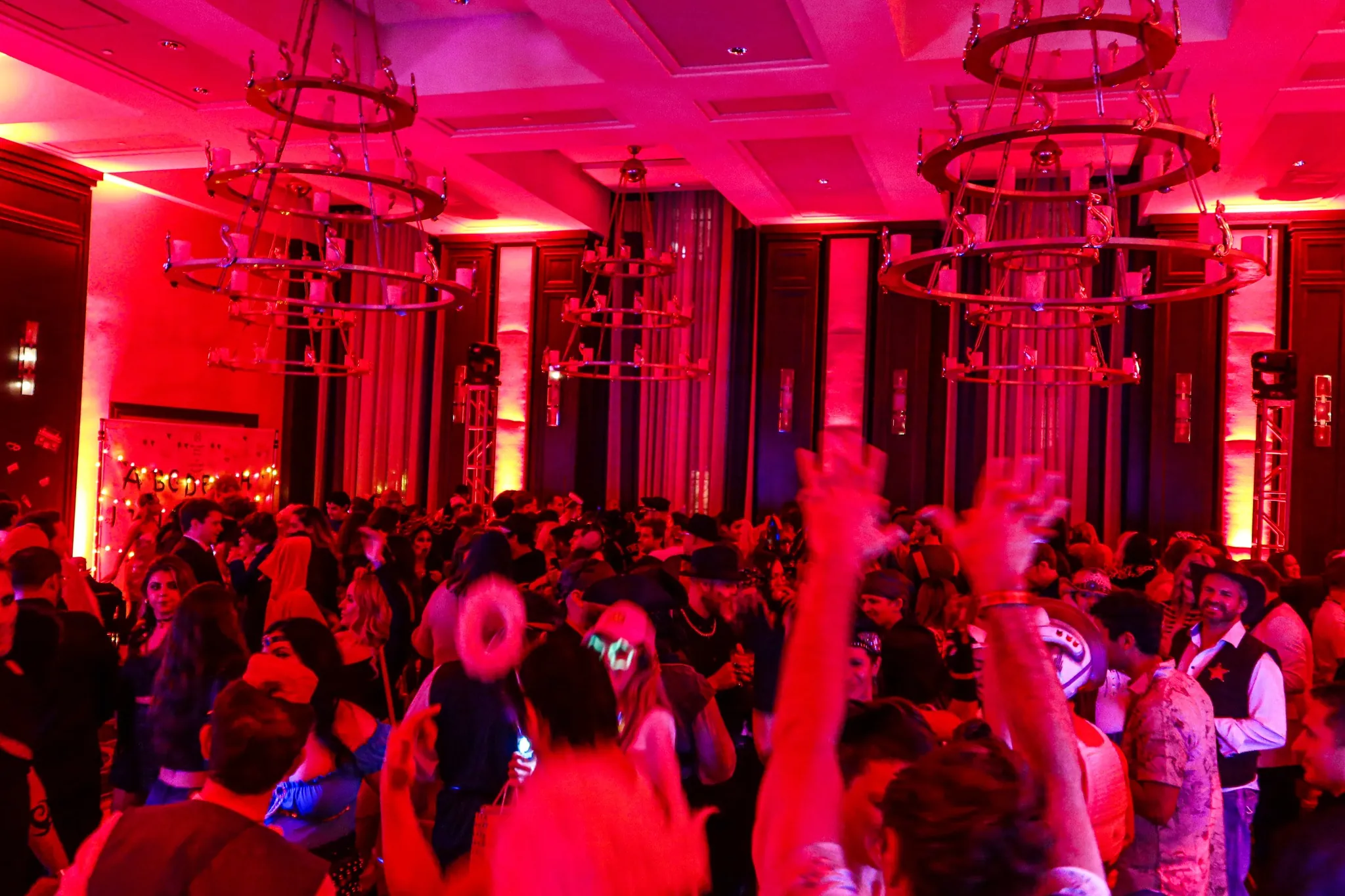 A large crowd of people dancing and celebrating at a lively indoor party with red lighting and chandeliers overhead.