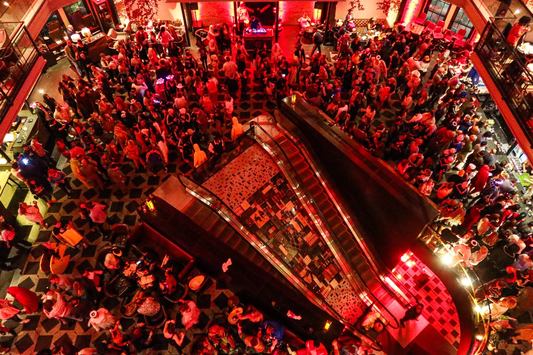 A large crowd gathers at a lively indoor event with red lighting, viewed from above near an escalator in a spacious venue.