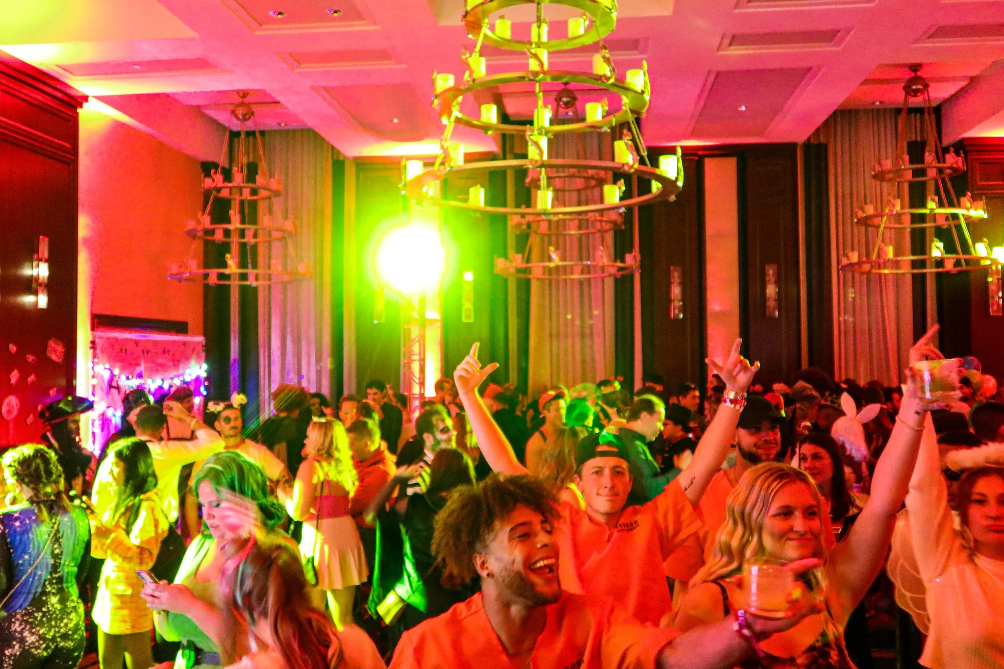 A lively crowd dances and celebrates at a brightly lit indoor party with colorful lights and decorations.