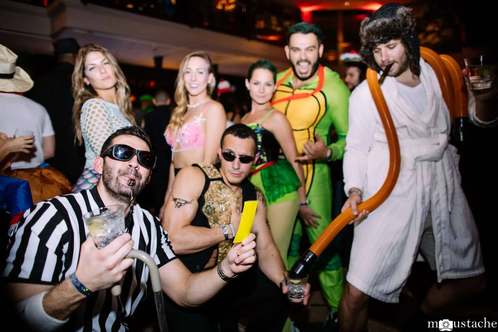 A group of people in costumes at a party: a referee, a wrestler, a Ninja Turtle, a man in a robe with tubes, and two women in festive outfits.