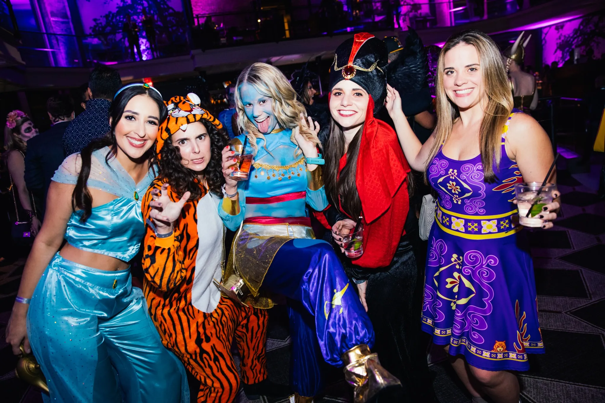 Five women in colorful costumes pose and smile at a lively party, holding drinks in their hands.