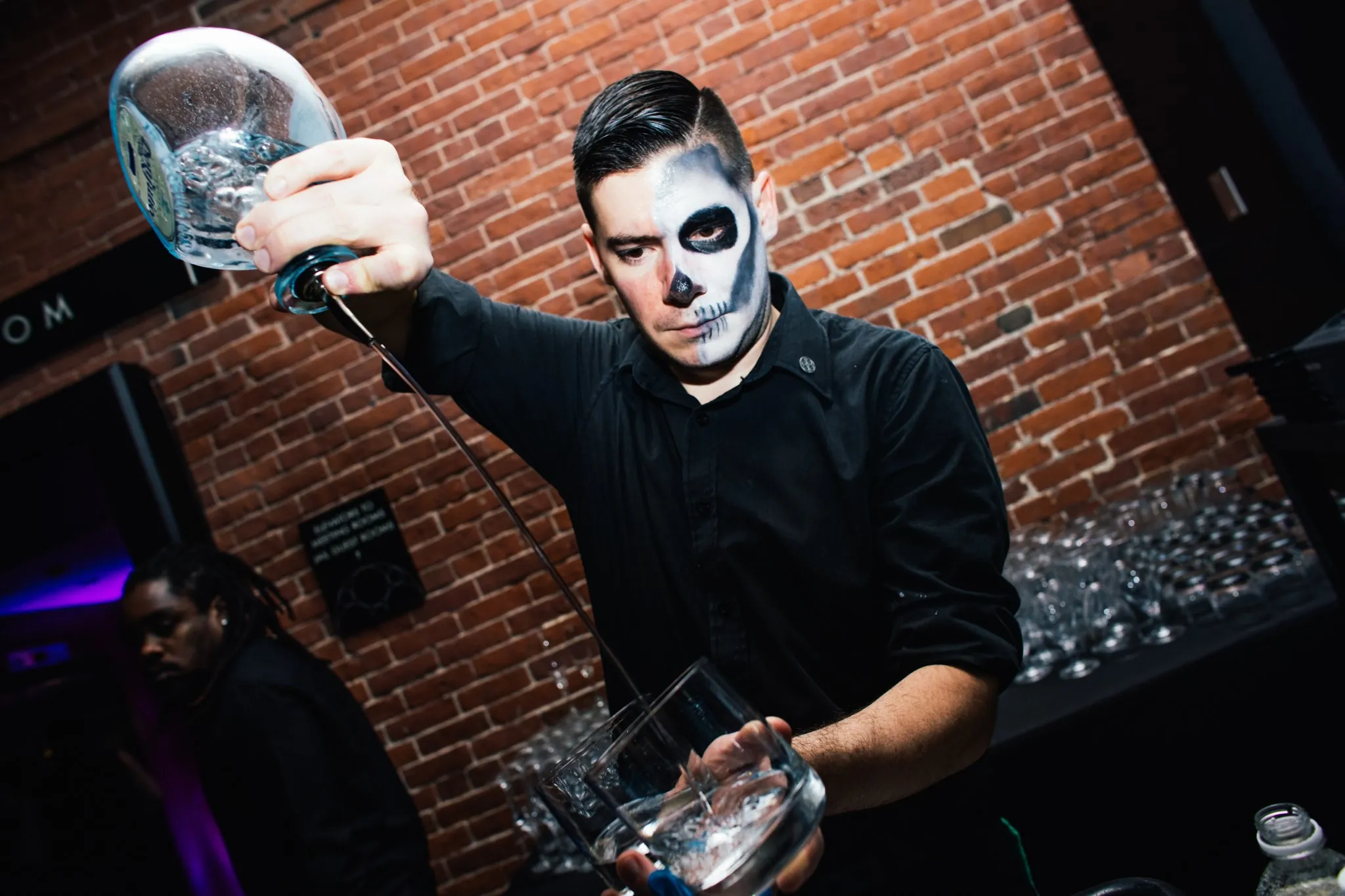 Bartender with half skull face paint pours liquor into a glass at a bar with a brick wall background.