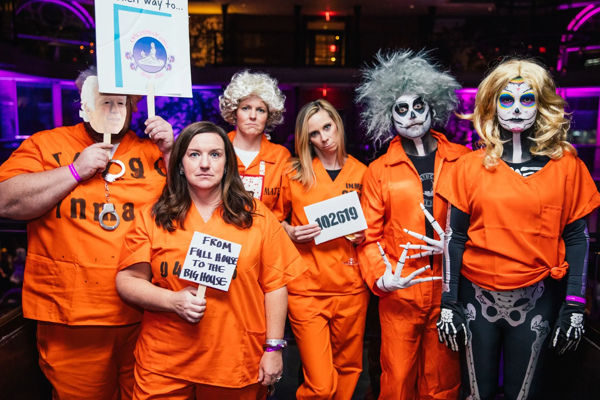 Six people in orange prison costumes, some with scary makeup and wigs, stand together at a party; one holds a sign and another holds a mugshot card.