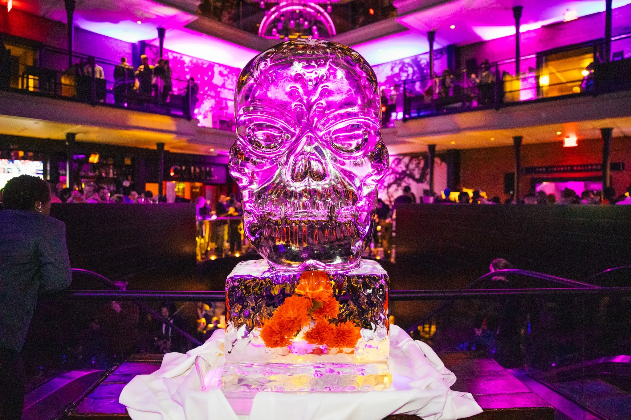 Large ice sculpture of a skull, illuminated with purple lighting, displayed at a crowded indoor event or party.