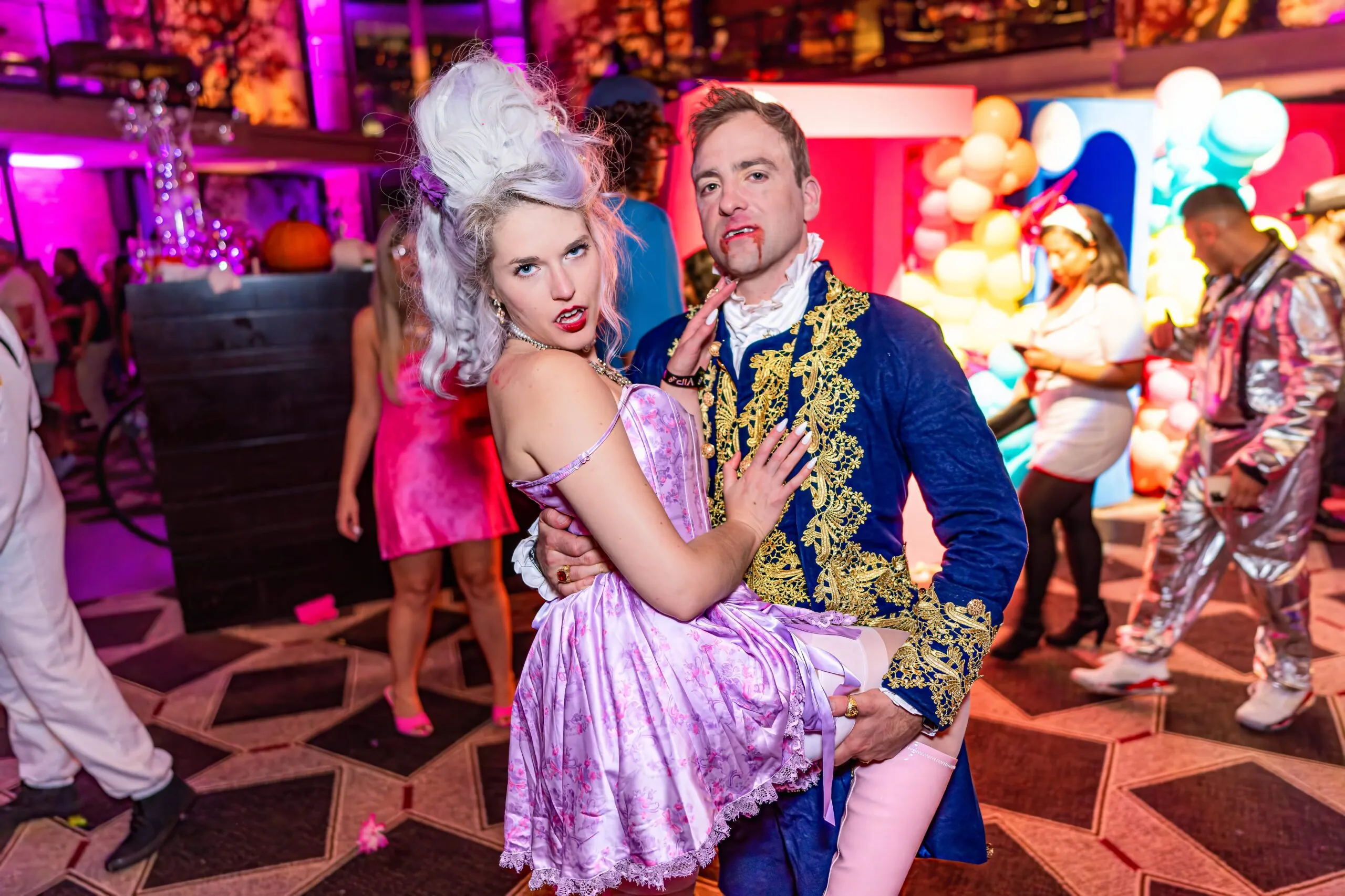 A woman in a pink Marie Antoinette costume is held by a man in a blue and gold period outfit with fake blood on his mouth at a lively costume party.