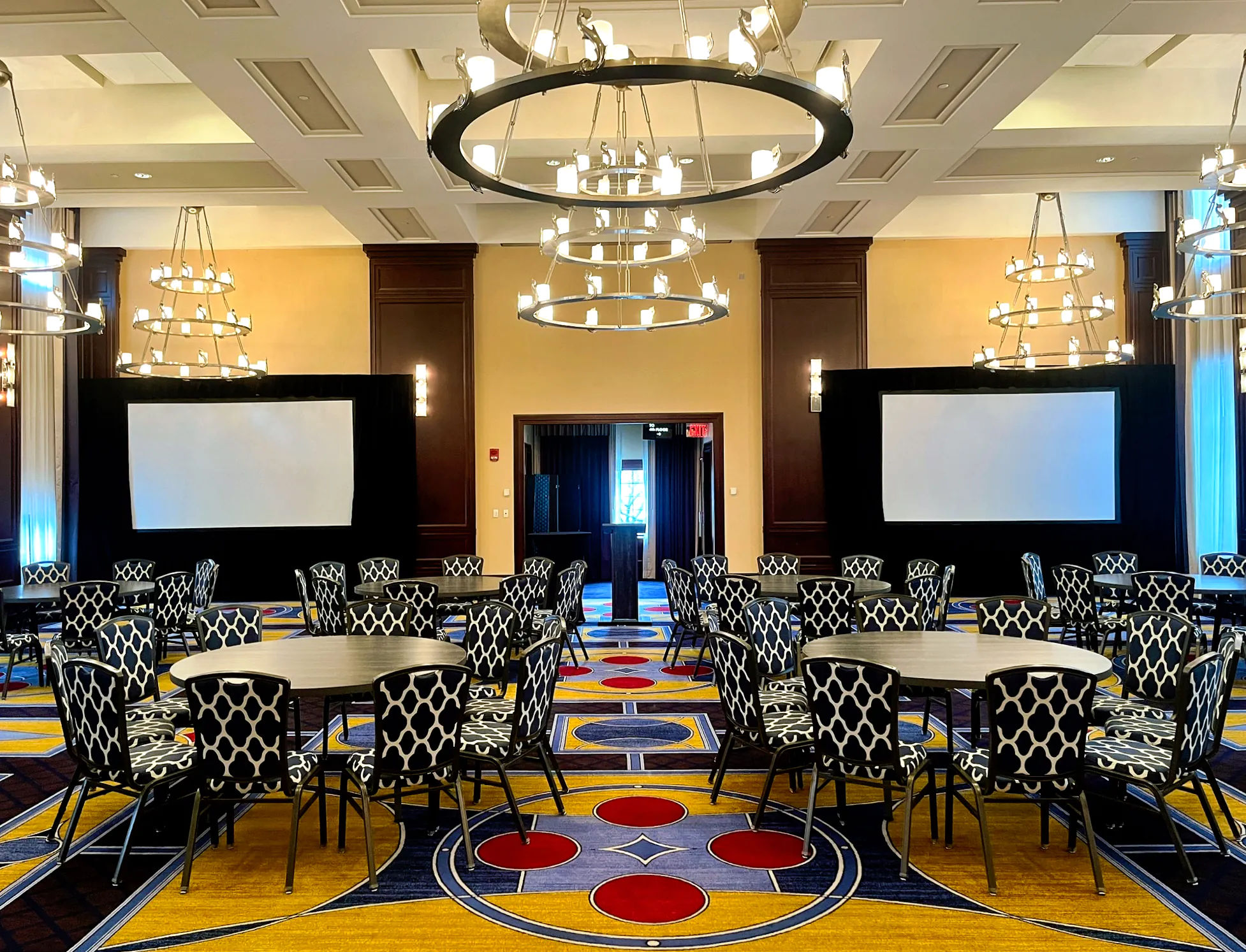 Conference room near Boston Common with round tables, patterned chairs, two large screens, and modern chandeliers hanging from the ceiling.